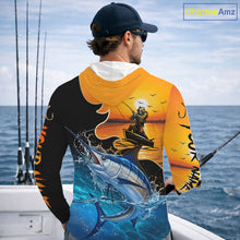 Load image into Gallery viewer, Person wearing a saltwater long sleeve hooded fishing shirt with a sunset and fish reaper design, standing on a boat.